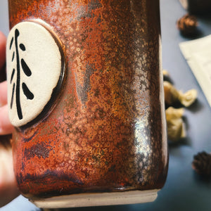 Brown ceramic mug with a circular emblem featuring a black symbol on a blurred natural background.