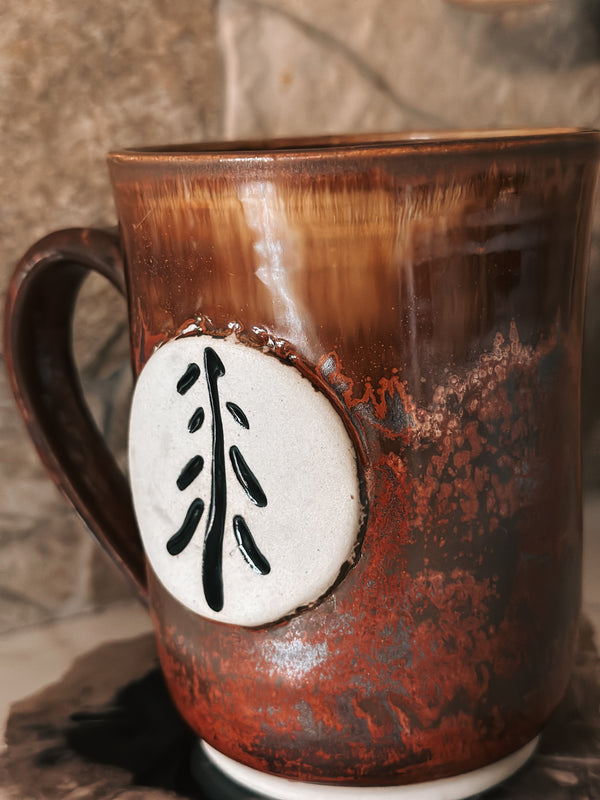 Brown ceramic mug with a white circular design featuring black leaf-like patterns on a stone surface.